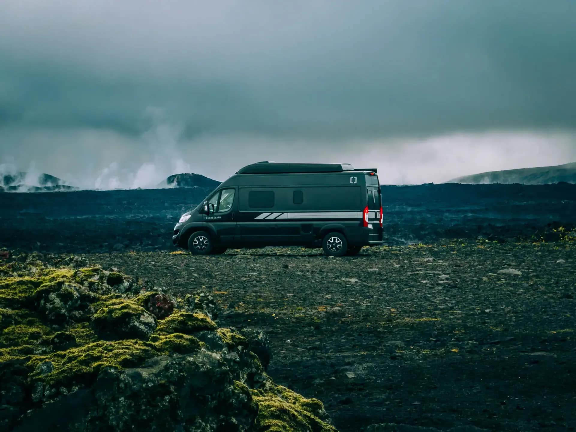 Black Crosscamp campervan parked on a rugged volcanic lava field in Iceland, with steam rising in the background under heavy dramatic clouds.