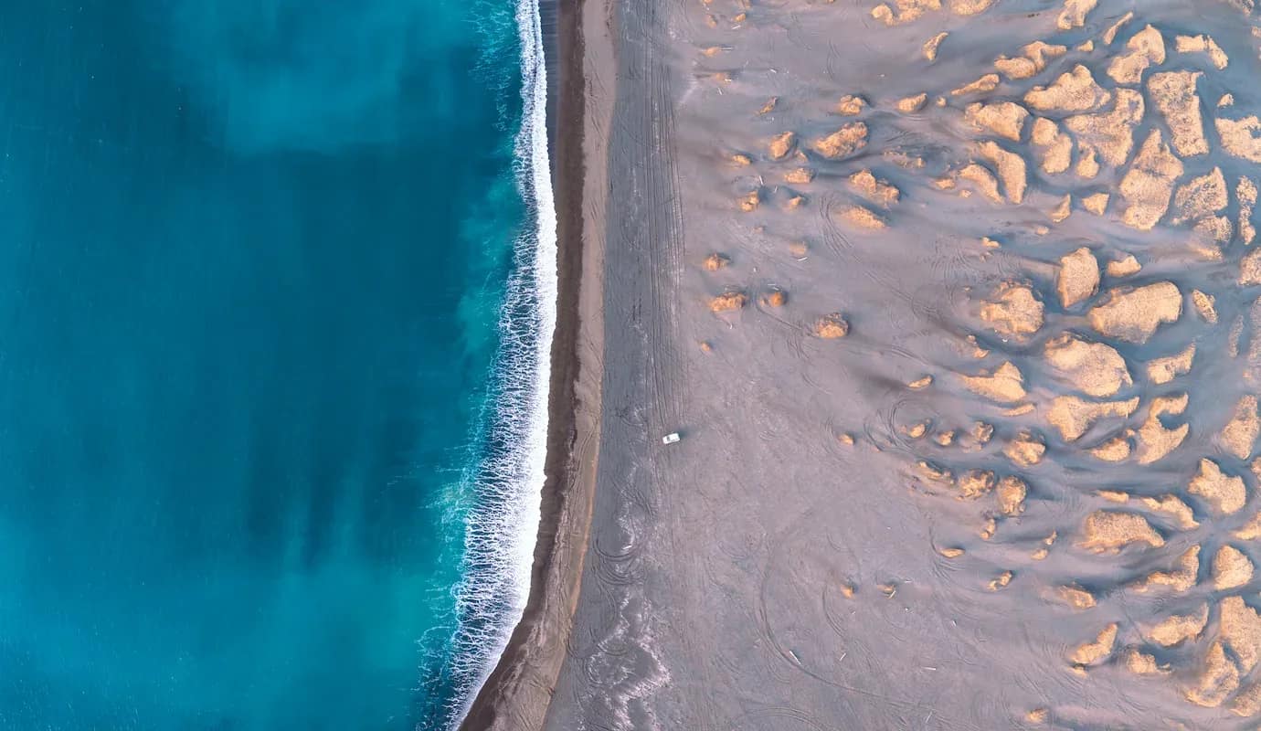 Aerial drone view of Iceland’s coastline showing turquoise ocean waves meeting a wide sandy beach with dune formations and a single vehicle parked near the shoreline.
