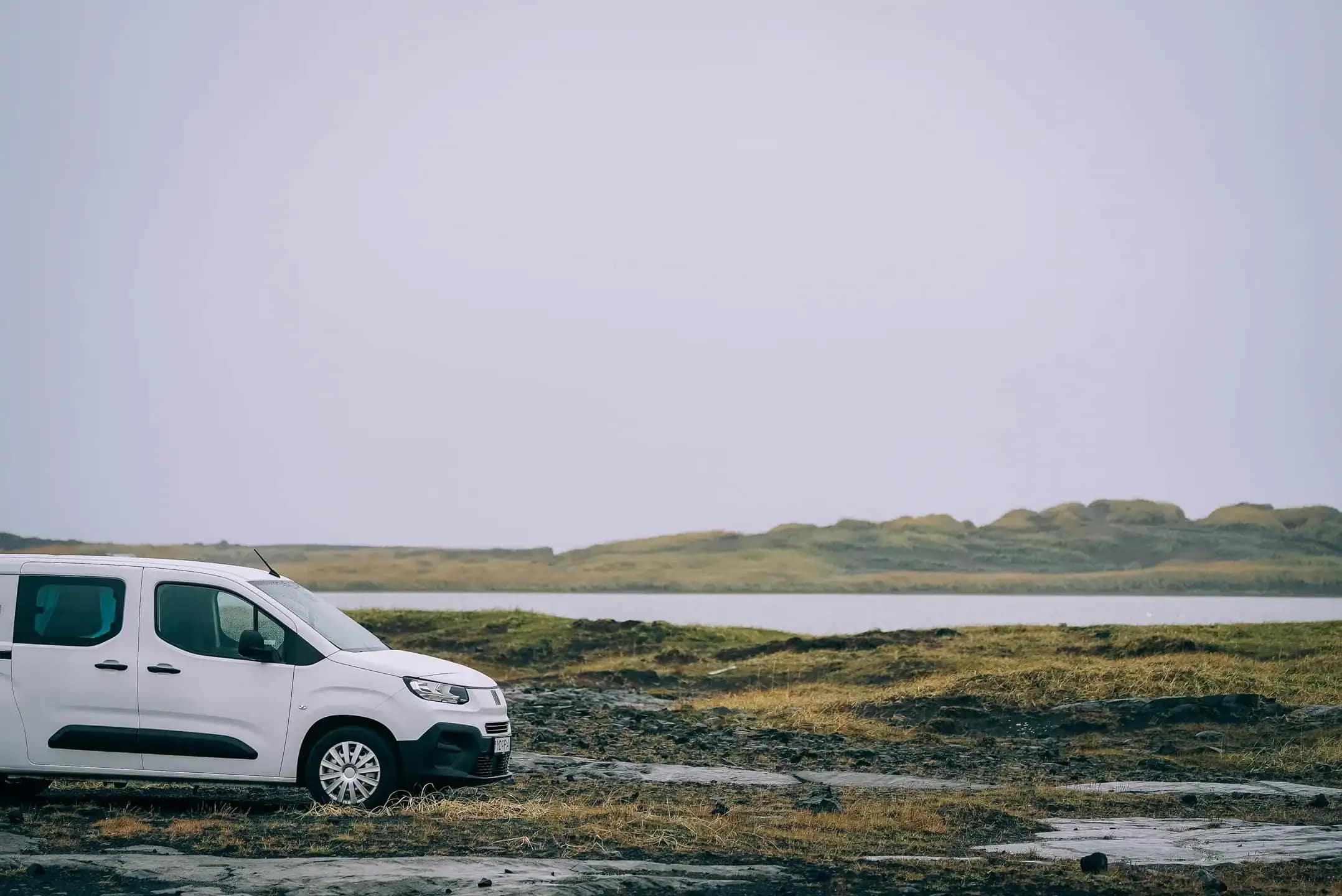 White Fiat Doblo campervan parked on a rocky moss landscape in Iceland beside a calm lake, with low green hills in the background under an overcast winter sky.