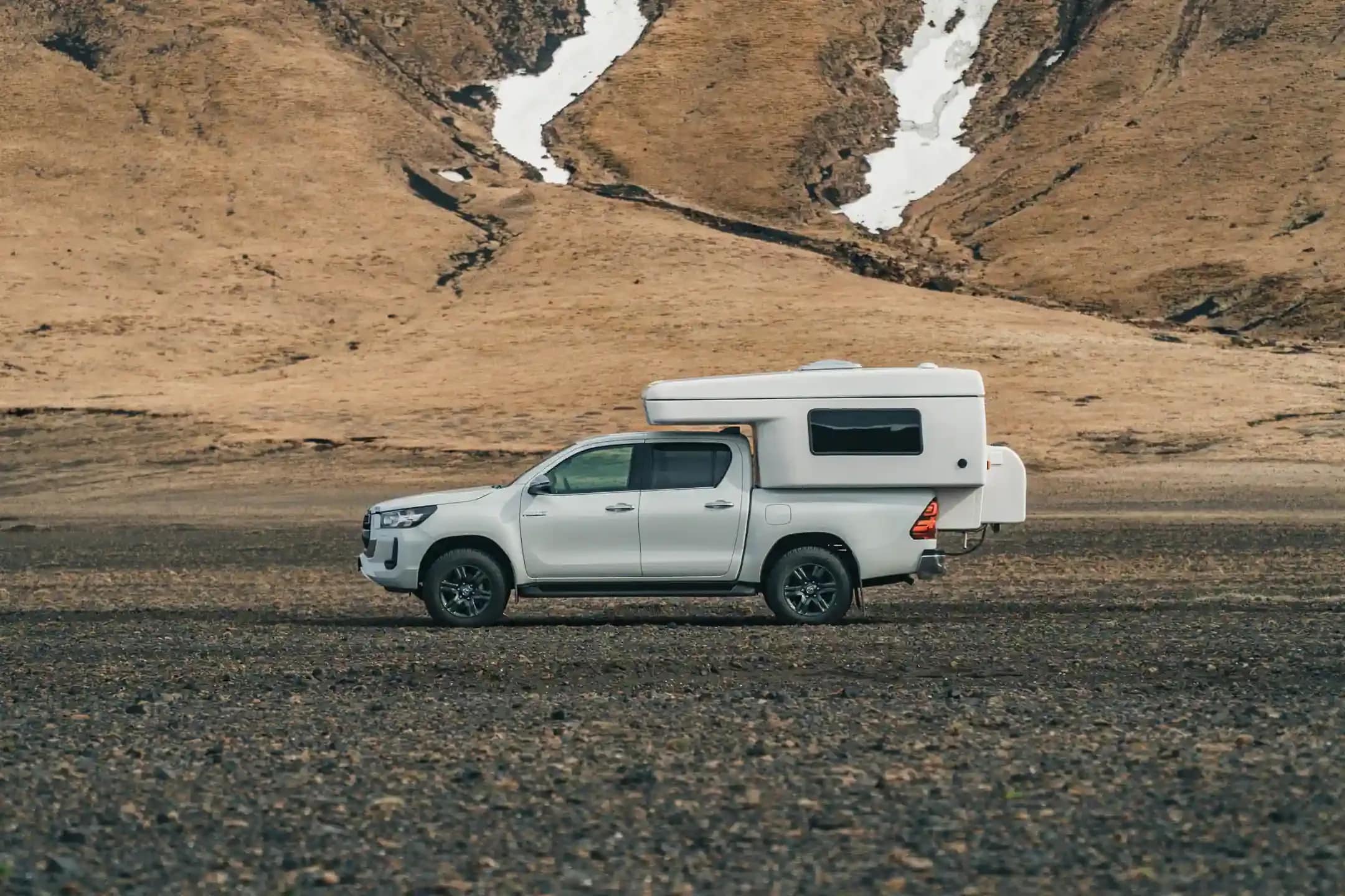 White Toyota Hilux 4x4 campervan parked on black sand in an Icelandic volcanic valley with snow patches on brown mountains