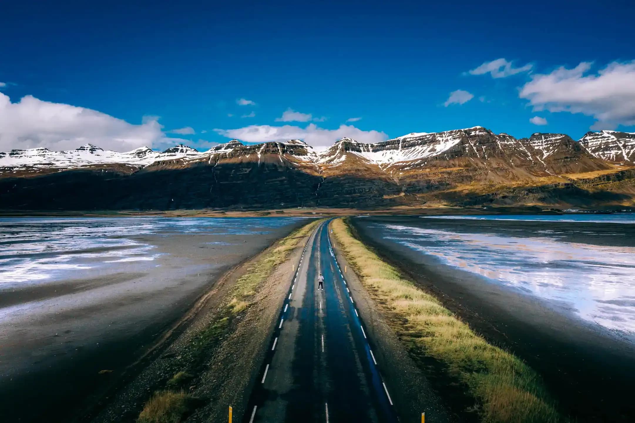 Straight Icelandic road stretching through a wide coastal plain with shallow water on both sides and snow capped mountains under a clear blue sky