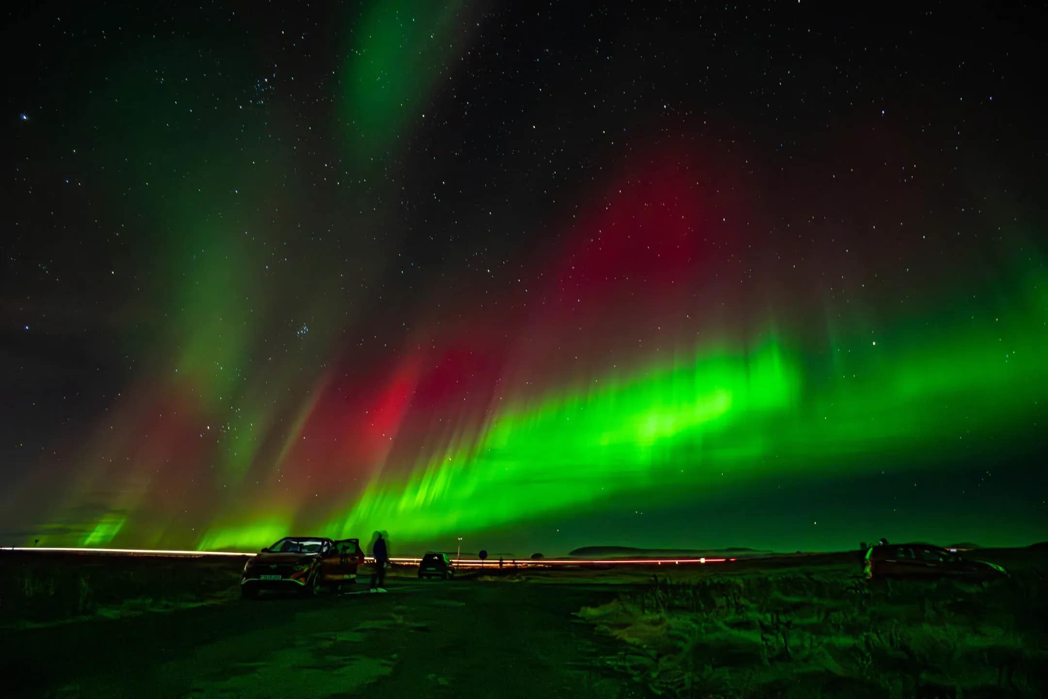 Bright green and red Northern Lights illuminating the night sky in Iceland with silhouettes of parked cars and travelers watching the aurora on a dark roadside