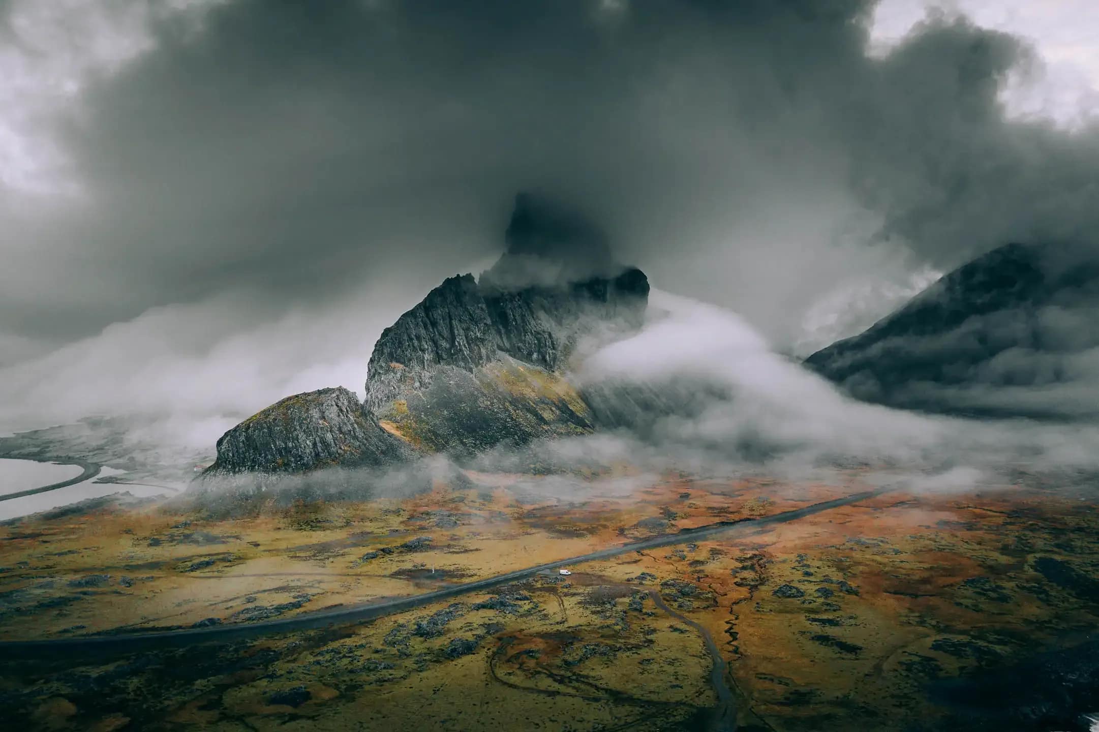 Aerial view of a winding road crossing a colorful volcanic plain with a small campervan beneath mist covered mountains in Iceland during a scenic road trip