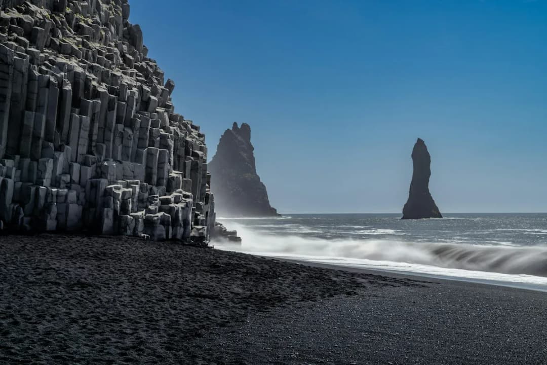 黑沙滩上的玄武岩石柱和远处的海蚀柱，海浪拍打着冰岛南岸的火山海岸