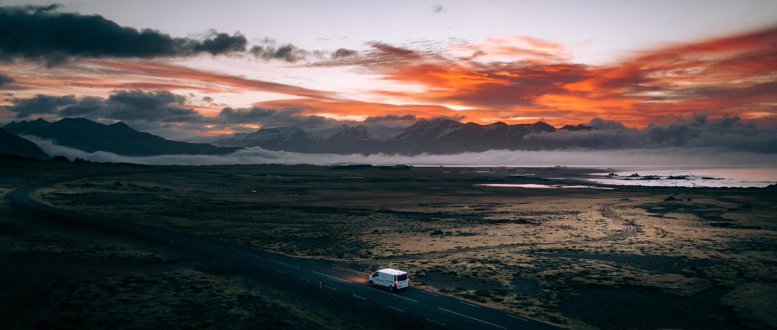 White Foxie campervan on an Icelandic road at sunset, with snow-capped mountains and a burning orange sky