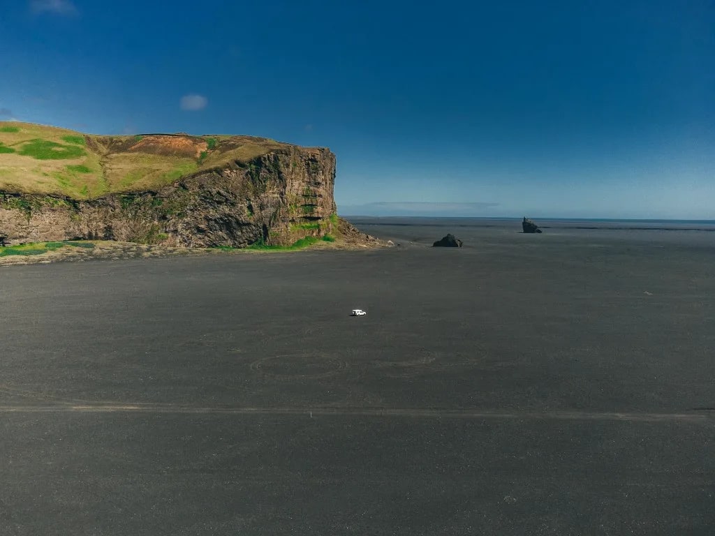 Aerial view of a white vehicle on a vast Icelandic black-sand flat beside a moss-topped cliff