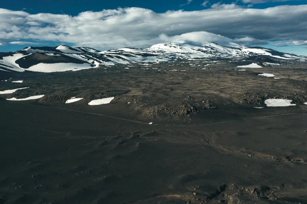 Aerial view of a lone white campervan on a highland track through dark volcanic rock and snow
