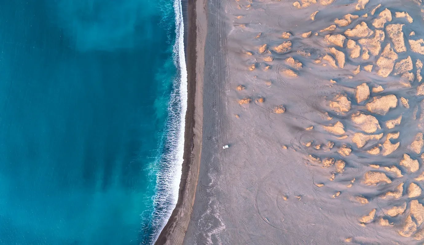 Aerial drone view of Iceland’s coastline showing turquoise ocean waves meeting a wide sandy beach with dune formations and a single vehicle parked near the shoreline.