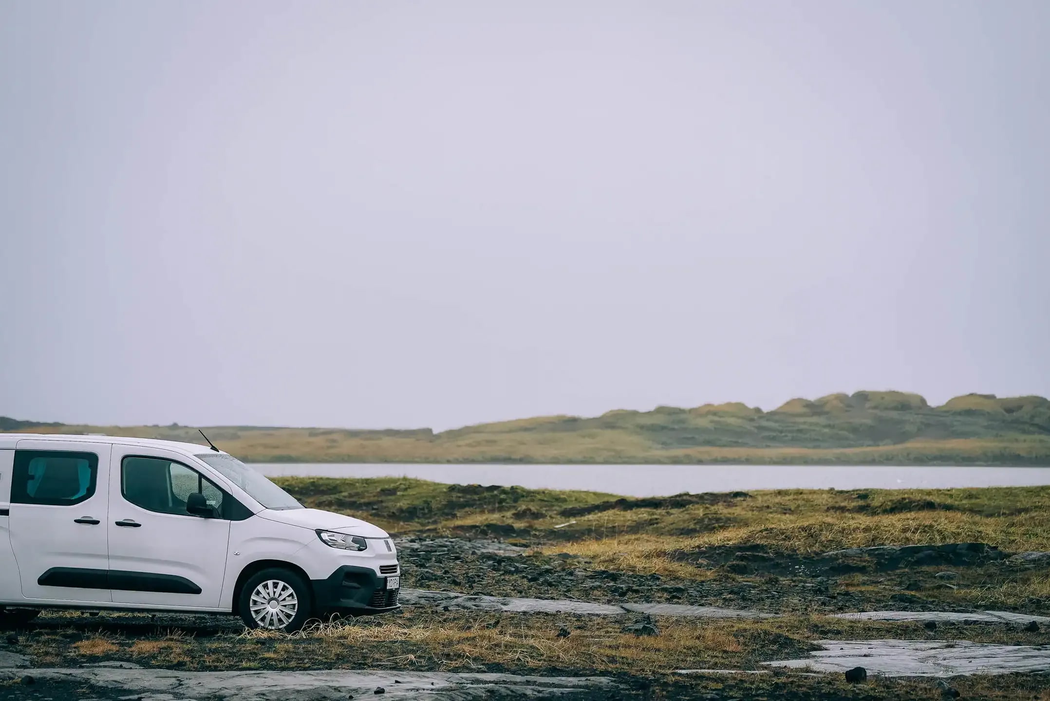 White Fiat Doblo campervan parked on a rocky moss landscape in Iceland beside a calm lake, with low green hills in the background under an overcast winter sky.