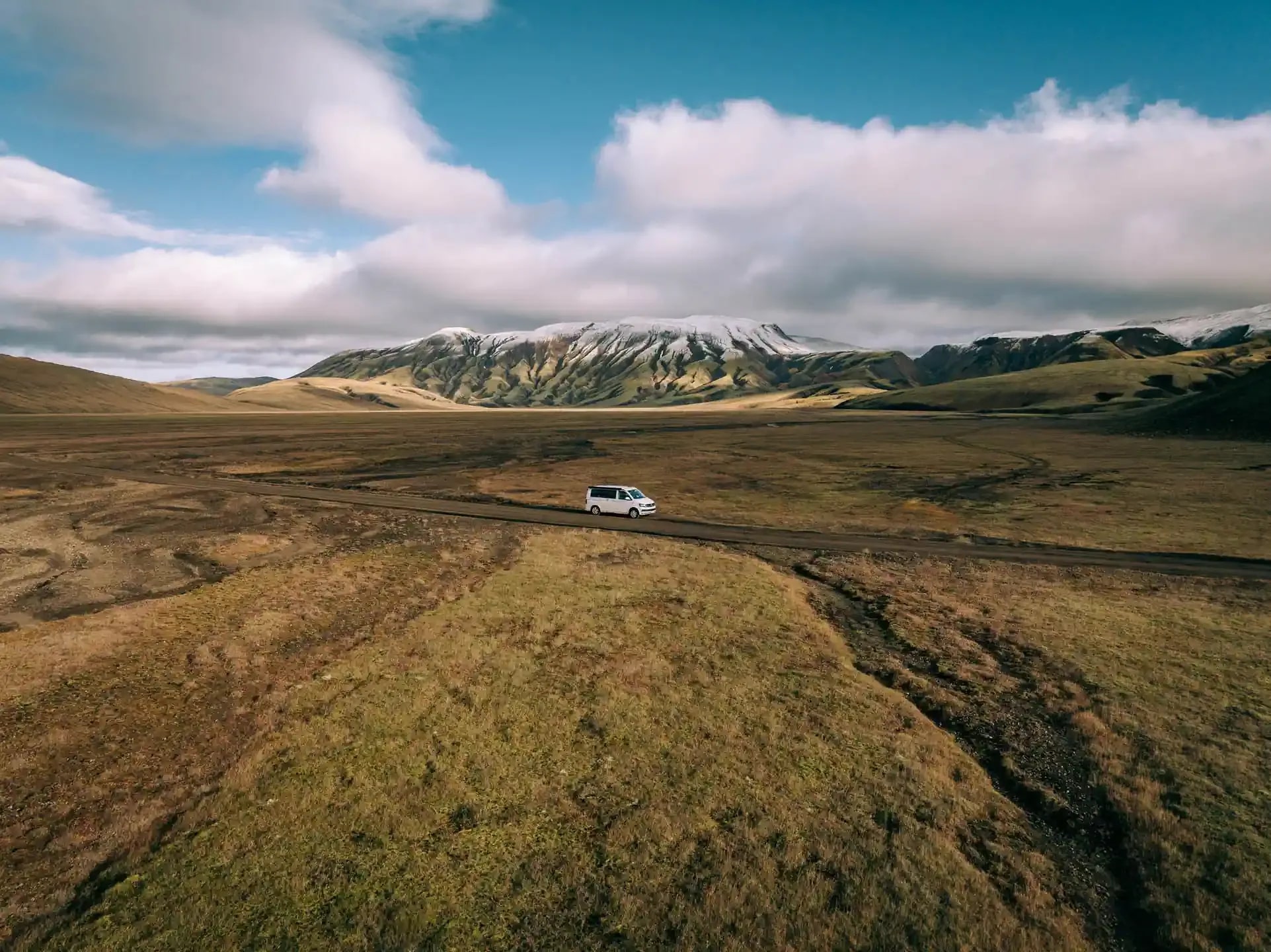 A white VW camper van driving along a dirt road through a vast, grassy plain with snow-capped mountains in the background under a cloudy blue sky