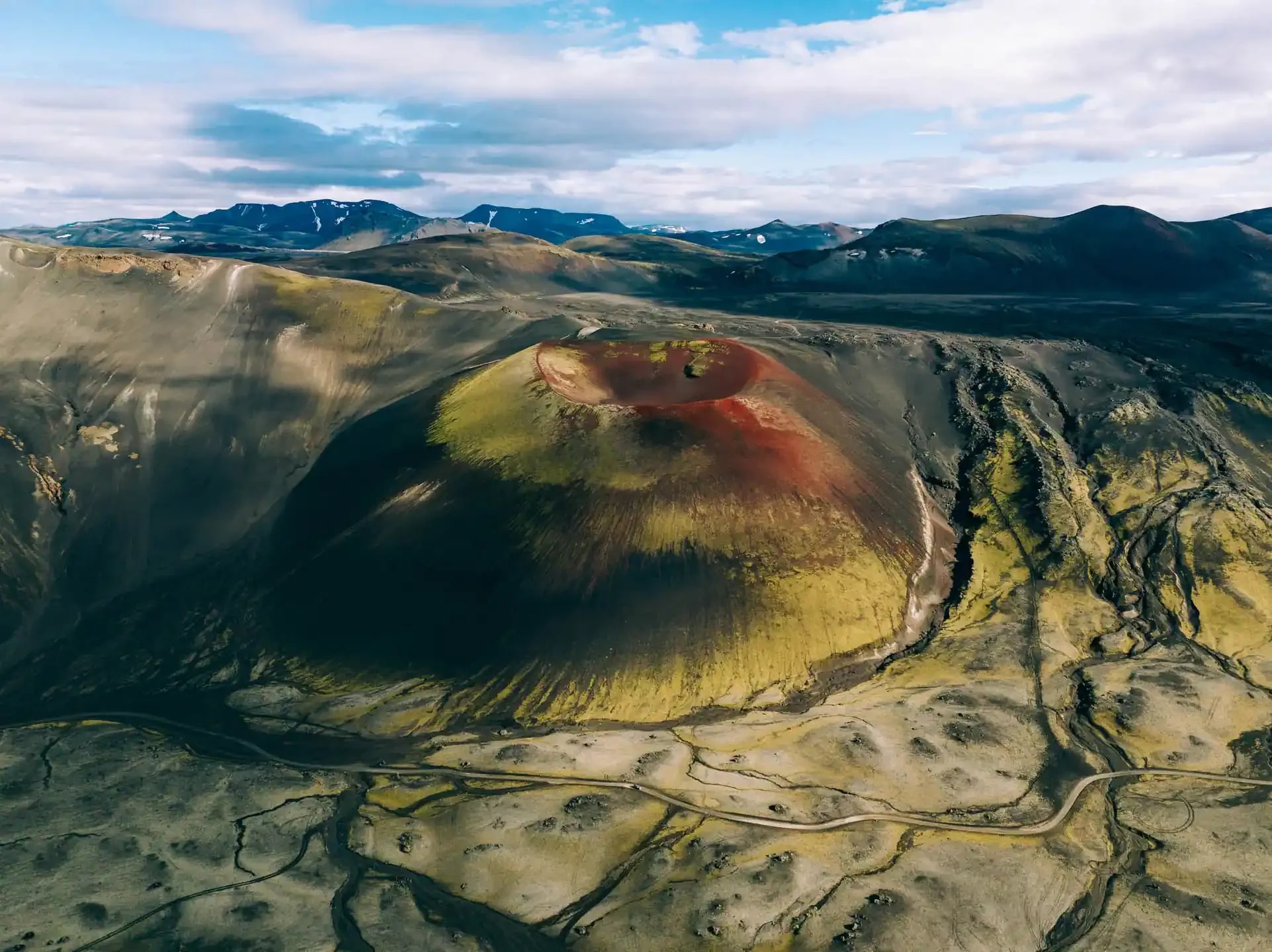A colorful volcanic crater in the Icelandic Highlands featuring striking red lava rock and green moss, with a remote gravel road winding through the volcanic landscape