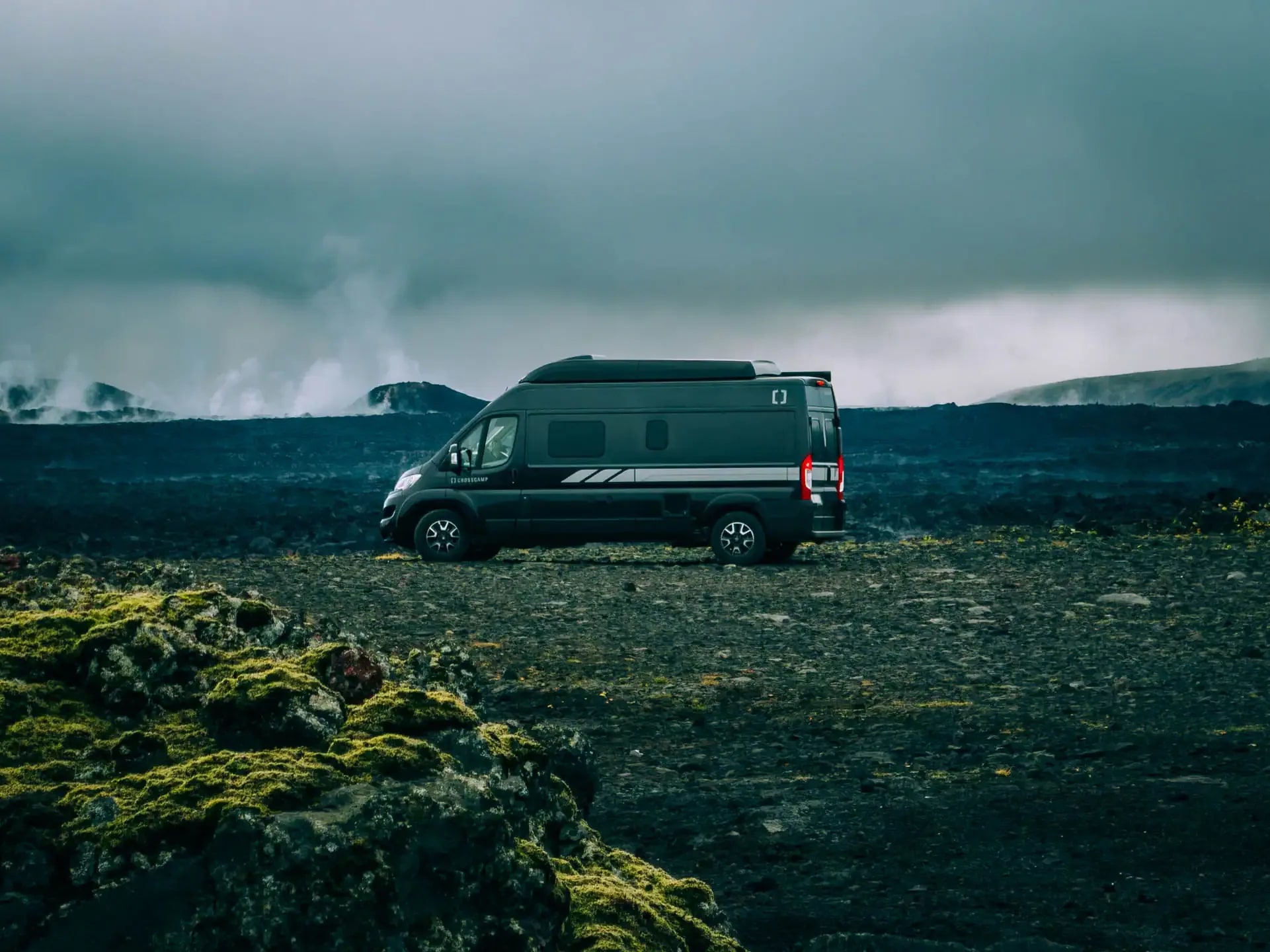Black Crosscamp campervan parked on a rugged volcanic lava field in Iceland, with steam rising in the background under heavy dramatic clouds.