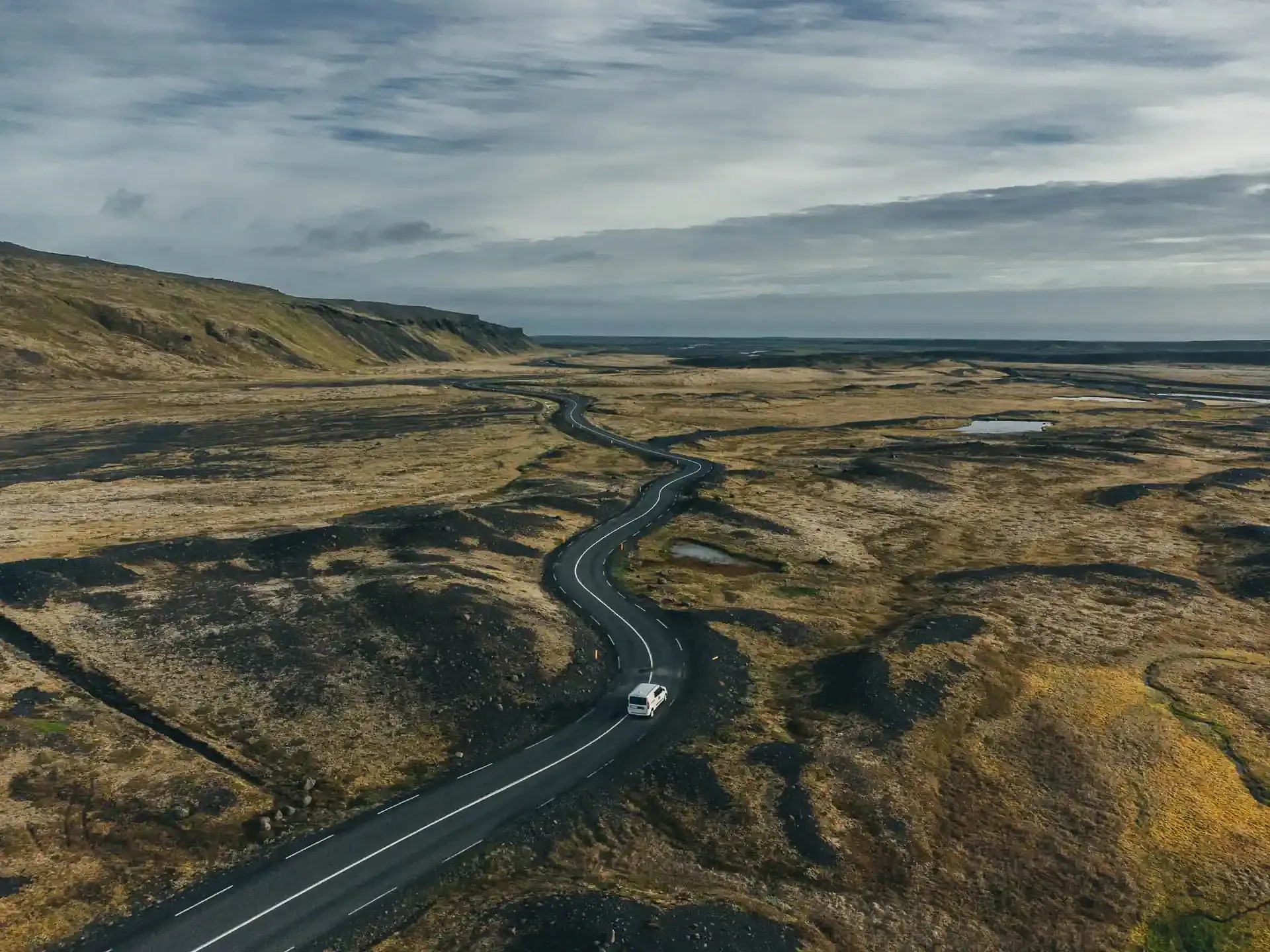 A white Fiat Doblo campervan driving along a winding paved road through a vast Icelandic landscape of mossy lava fields and rolling hills under a cloudy sky