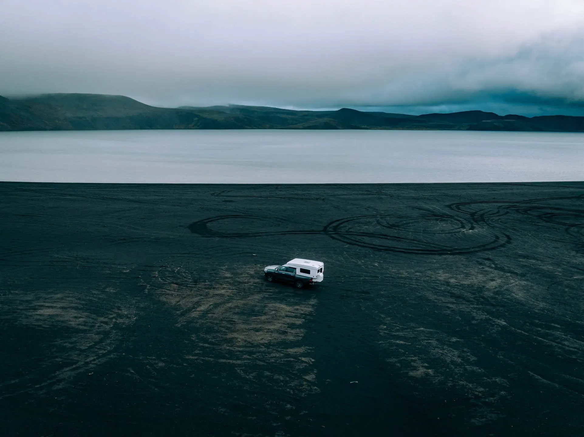 4x4 pickup camper driving across a black volcanic beach beside a calm lake with distant mountains under an overcast sky in Iceland