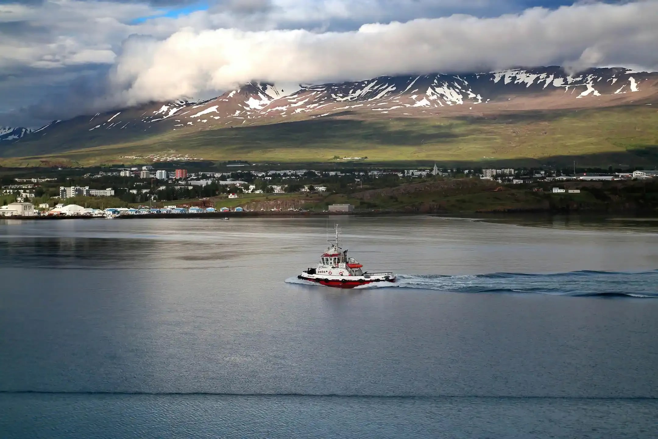 Red and white boat sailing across Akureyri harbor in North Iceland with snow-capped mountains and green hills in the background