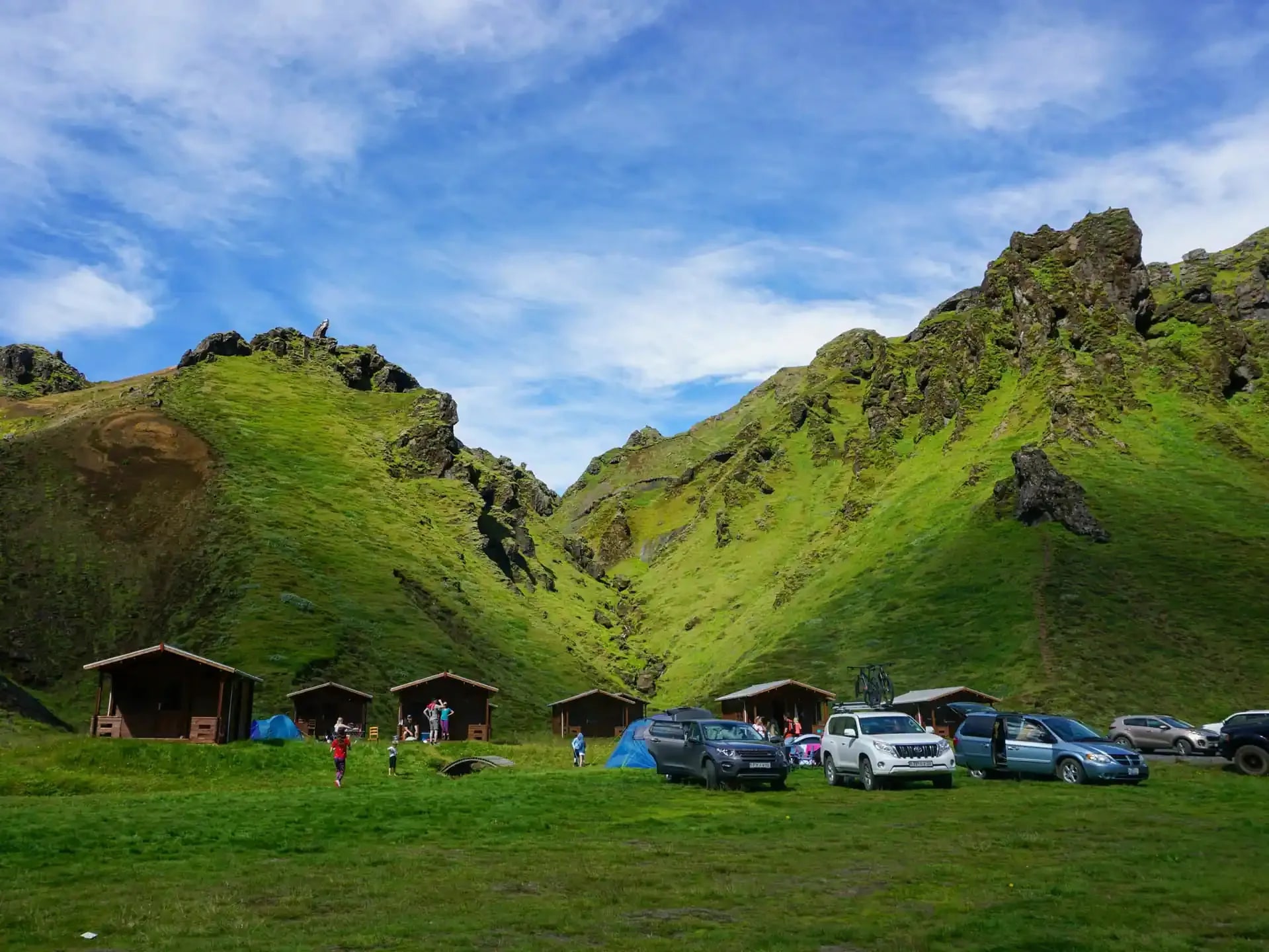 Cars, campers, and wooden cabins in lush green Icelandic mountain campsite under blue spring sky