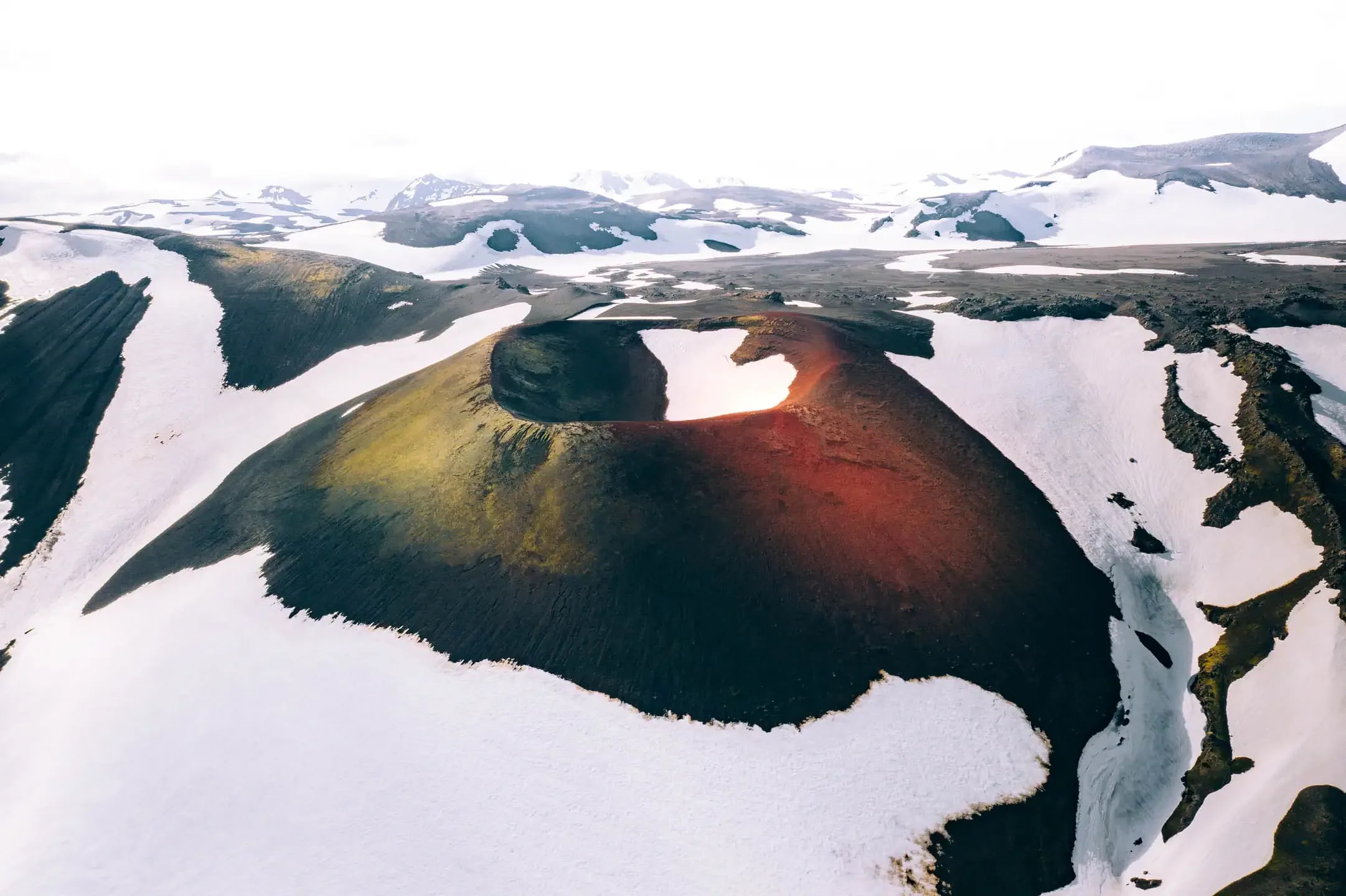 Aerial view of a volcanic crater surrounded by black lava fields and patches of melting snow in the Icelandic highlands