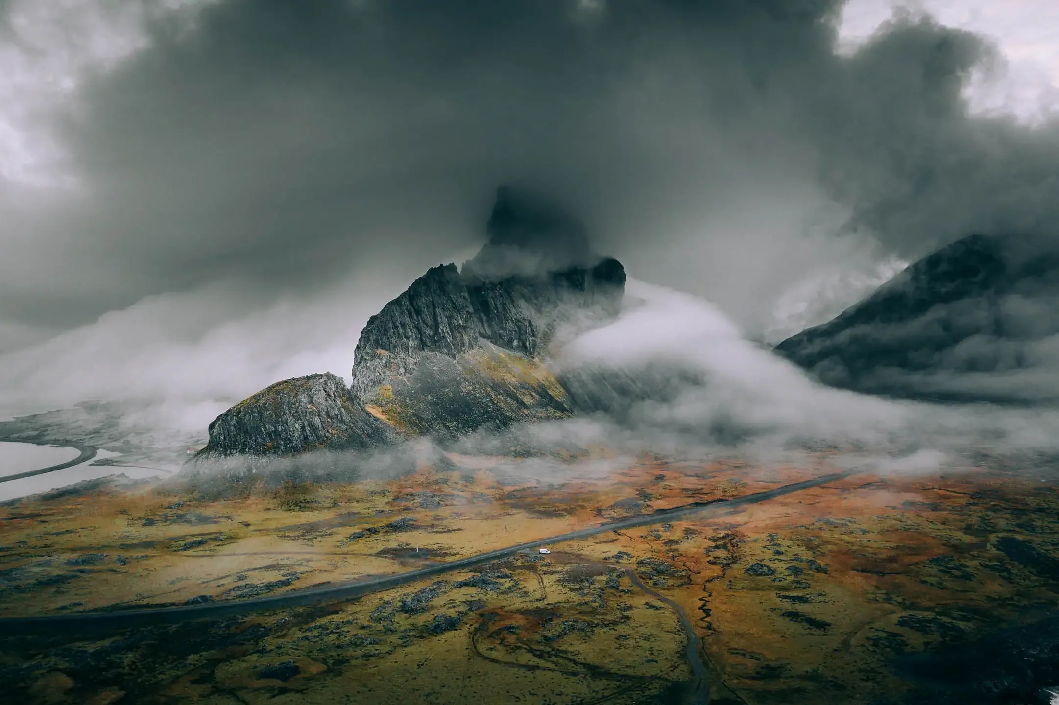 Aerial view of a winding road crossing a colorful volcanic plain with a small campervan beneath mist covered mountains in Iceland during a scenic road trip